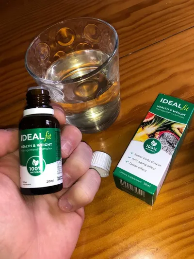 Mano que sostiene el frasco de IdealFit junto a un vaso de agua y la caja